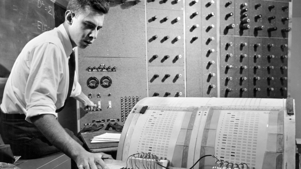 Investigador traballando nunha computadora de primeiros tempos, anos cincuenta do século XX. Ao fondo, panel de conexións electrónicas.